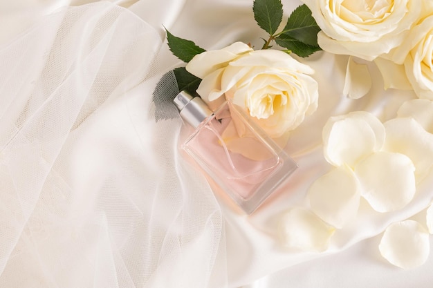 A transparent bottle of women's perfume or spray lies on a chic satin fabric creamy background among roses and petals Advertising concept