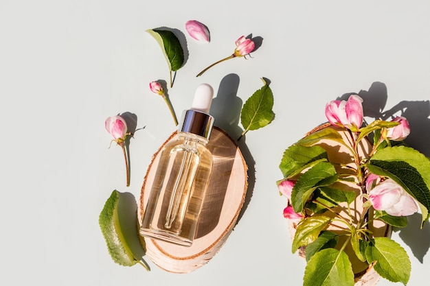 A transparent cosmetic bottle with a natural face and body care product lies on a wooden tree slice among the spring flowers of an apple tree