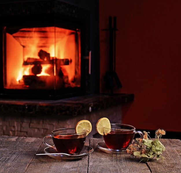 Transparent cups of hot tea with lemon and dry linden over\
burning fireplace on wooden table valentine winter holiday\
concept