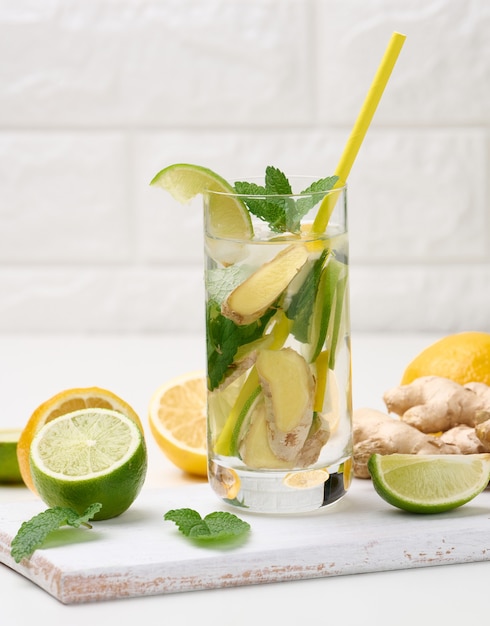 Tazza di vetro trasparente con limone, pezzi di lime e foglie di menta e pezzi di zenzero su un tavolo bianco, detox.