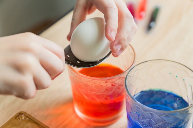 Transparent glass cups with colored water. child hands put in a white egg with a spoon.