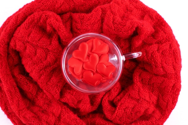 A transparent heart-shaped cup with red hearts inside stands on a red knitted scarf. The Concept Of Valentine's Day. Flat lay.