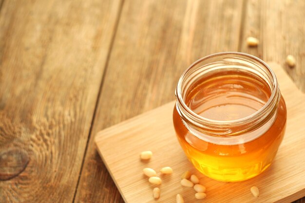 Photo a transparent jar of honey stands on a board on a wooden table nearby are pine nuts copy space selective focus