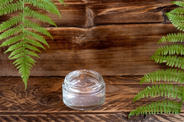 Transparent jar of moisturizer gel cream on wooden board with fern leaves. SPA natural organic beauty product packaging design.