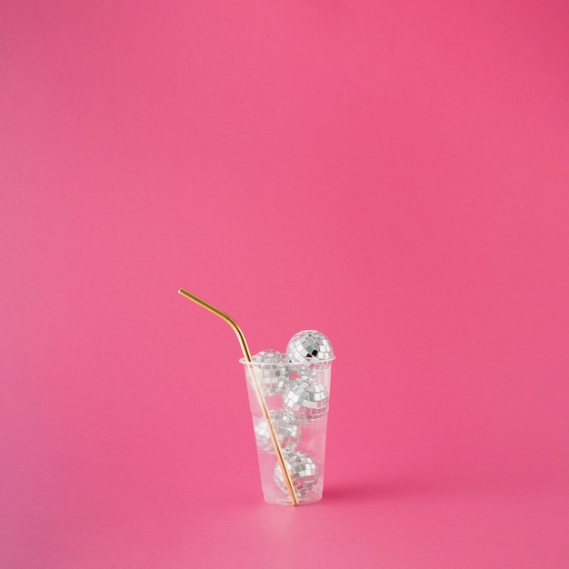 Transparent plastic drinking cup filled with shiny disco balls and golden straw on pink background