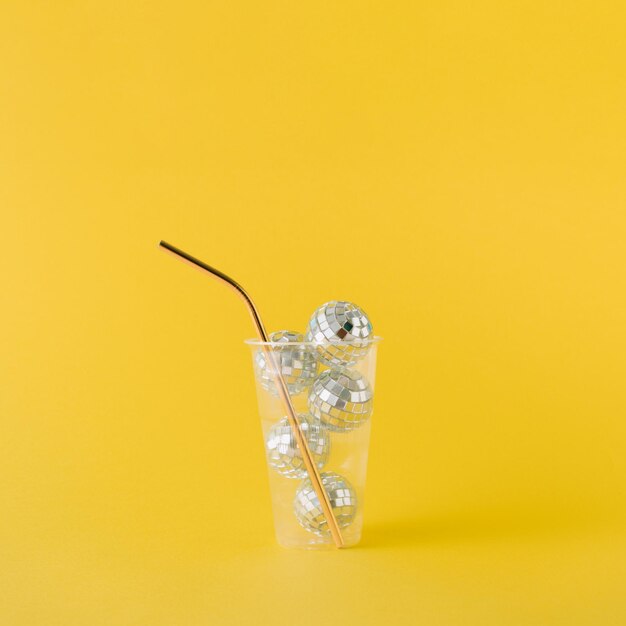 Transparent plastic drinking cup filled with shiny disco balls and golden straw on yellow background