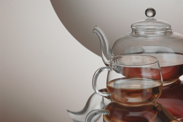 Transparent teapot with teacup on the reflective surface on a light gray background