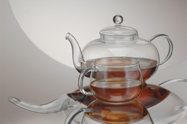 Transparent teapot with teacup on the reflective surface on a light gray background