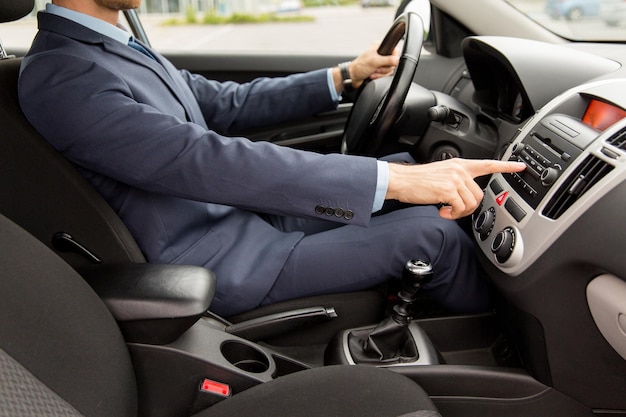 transport, business trip, technology and people concept - close up of young man in suit driving car and adjusting music volume on control panel stereo system