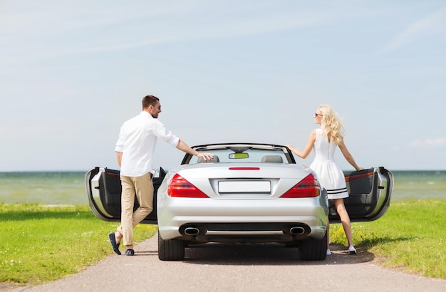 transport, travel, road trip and people concept - happy man and woman near cabriolet car at sea side