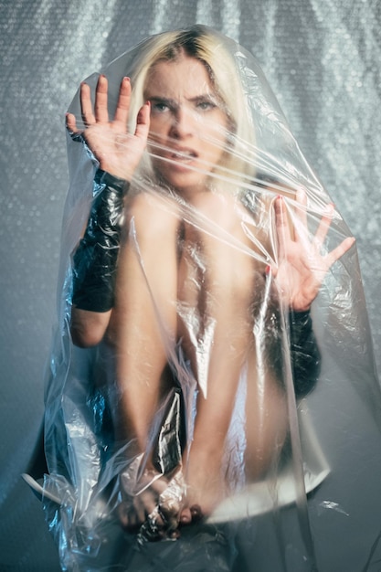 Trapped woman. Defocused art portrait. Depression self-isolation. Scared helpless lady on chair looking through covered transparent plastic film isolated on gray blur.