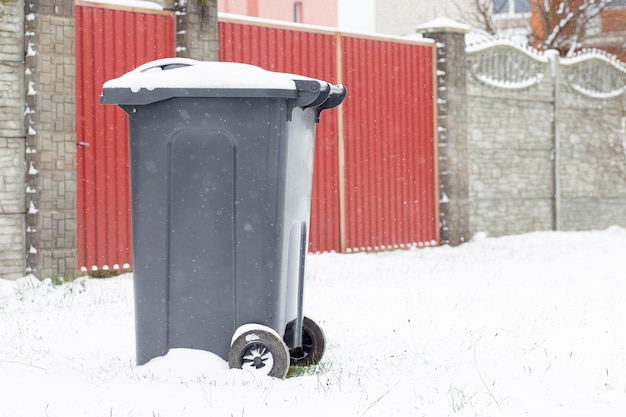 A trash can covered with snow next to a house on the street Garbage removal in winter