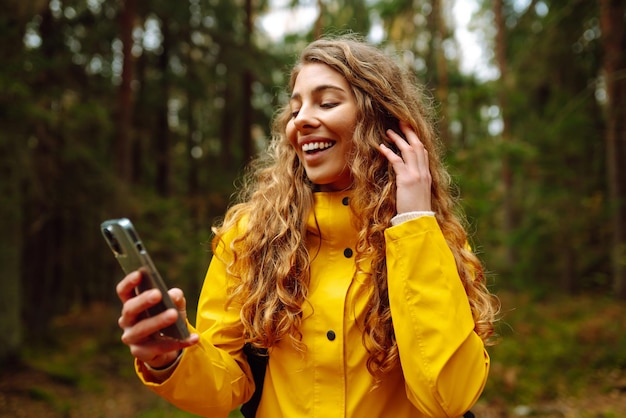 Приложения для путешествий Кудрявая молодая женщина-путешественница в желтом пальто держит в руке смартфон Концепция путешествия