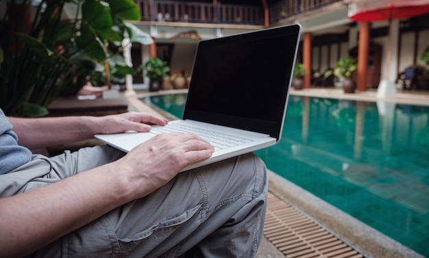 Travel blogger sitting at swiming pool writing article on white laptop