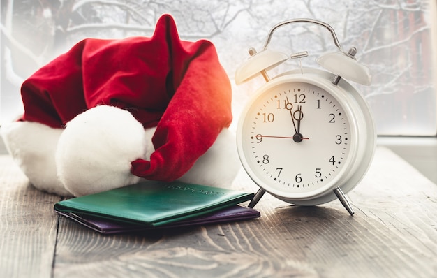 Travel on christmas concept. Vintage alarm clock with santa claus hat and passport on wooden table against the window