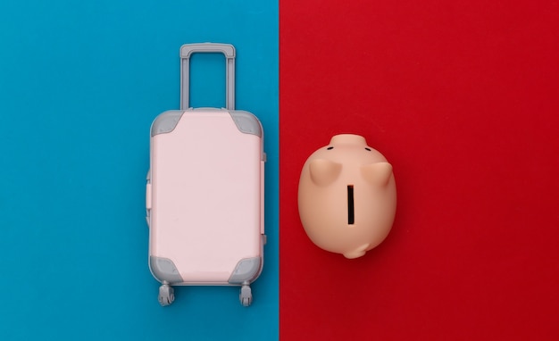 Travel concept. Mini plastic travel suitcase, piggy bank on red blue background. Minimal style. Top view. Flat lay