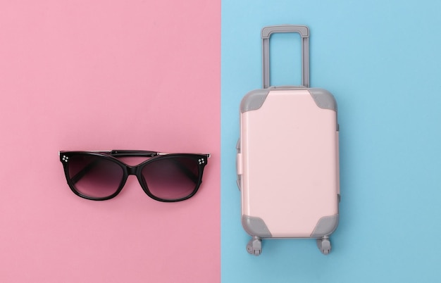 Travel concept. Mini plastic travel suitcase and sunglasses on pink blue pastel background. Minimal style. Top view, flat lay