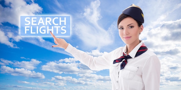 Travel concept - stewardess in uniform pointing at search flights button over blue sky background