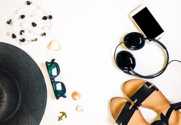 Foto viaggio accessori femminili telefono, auricolari, occhiali da sole, sandali, collana e cappello su bianco.