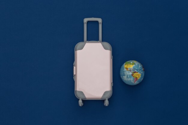 Travel flat lay. Mini plastic travel suitcase, globe on classic blue background. Minimal style. Top view