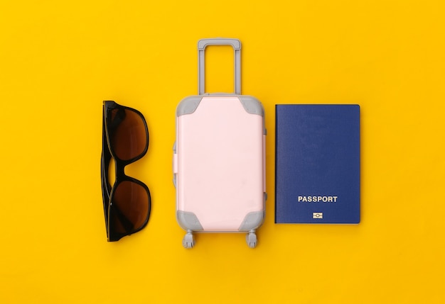 Travel flat lay. mini plastic travel suitcase, sunglasses and passport on yellow background. minimal style. top view