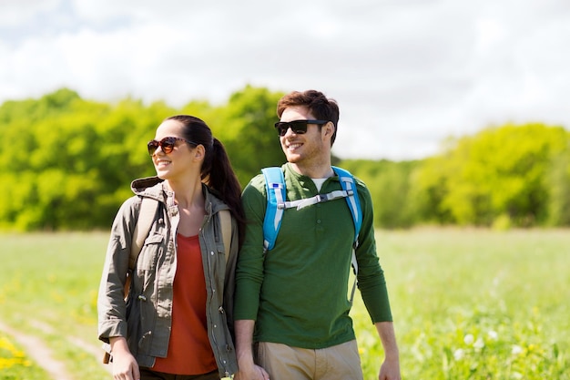 travel, hiking, backpacking, tourism and people concept - happy couple with backpacks holding hands and walking along country road outdoors