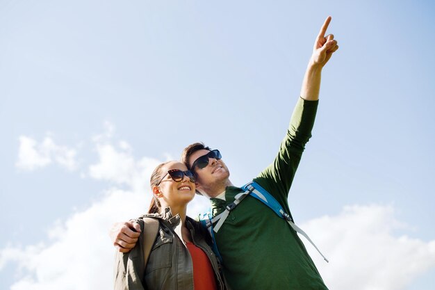 travel, hiking, backpacking, tourism and people concept - happy couple with backpacks outdoors pointing finger to something in sky