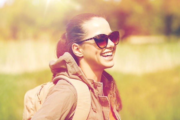 travel, hiking, backpacking, tourism and people concept - happy young woman in sunglasses with backpack walking along country road outdoors