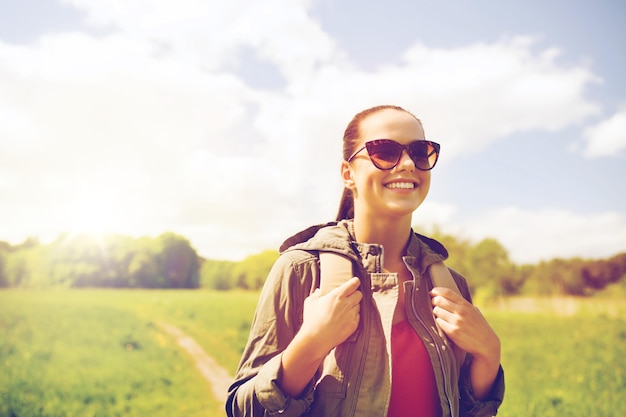 여행, 하이킹, 배낭여행, 관광, 그리고 사람들의 개념 - 야외에서 시골길을 따라 배낭을 메고 선글라스를 쓴 행복한 젊은 여성
