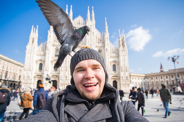 旅行、休日、冬の休暇のコンセプト-幸せな男はドゥオーモミラノ大聖堂の前で面白い鳩と一緒に自分撮り写真を撮ります