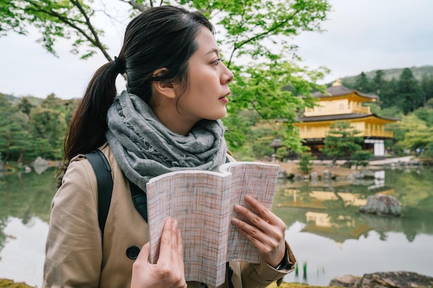 京都旅行。ガイドブックを持って日本の黄金寺院を背景に道を探す女性観光客。旅行者のライフ スタイル。