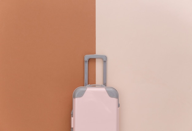 旅行のミニマリズム。ベージュブラウンの背景にミニプラスチック製の旅行スーツケース。最小限のスタイル。上面図、フラットレイ