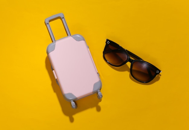 旅行のミニマリズム。深い影と黄色の背景にミニプラスチック製の旅行スーツケースとサングラス。最小限のスタイル。上面図、フラットレイ