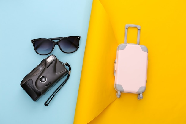 Планирование путешествия. Игрушка дорожный багаж, солнцезащитные очки и фотоаппарат на желтой синей бумаге. Плоская планировка