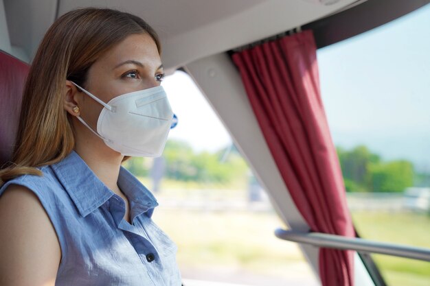 대중교통으로 안전하게 여행하세요. KN95 FFP2 보호용 안면 마스크를 쓴 젊은 여성이 여행 중 버스 창문을 통해 바라보고 있습니다.