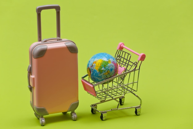 Travel and shopping concept. Mini plastic travel suitcase and supermarket trolley with globe on green background.