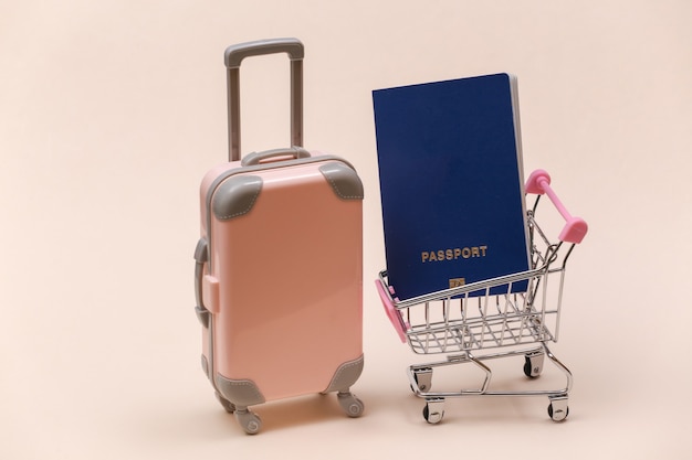 Travel and shopping concept. Mini plastic travel suitcase and supermarket trolley with passport on beige background.