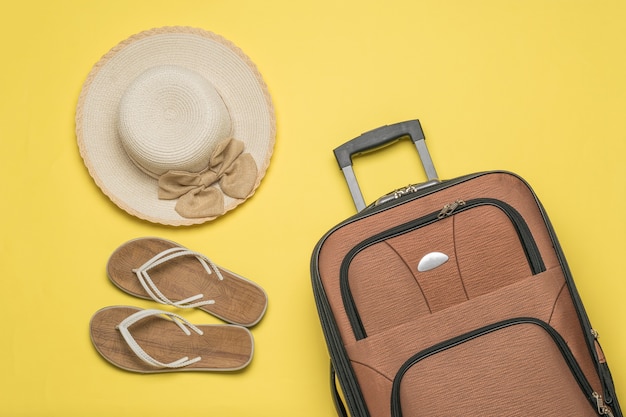 A travel suitcase, a woman's hat and sandals on a yellow background. Flat lay.