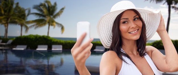 travel, summer, technology and people concept - sexy young woman taking selfie with smartphone over resort beach with palms and swimming pool background