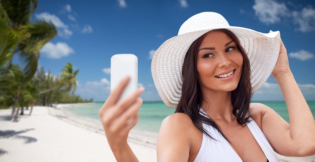 旅行、夏、技術、人々 のコンセプト - 手のひらの背景を持つ熱帯のビーチでスマート フォンで selfie を取るセクシーな若い女性