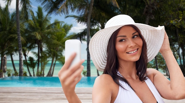 여행, 여름, 기술, 그리고 사람들의 개념 - 야자수와 수영장을 배경으로 열대 해변에서 스마트폰으로 셀카를 찍는 섹시한 젊은 여성