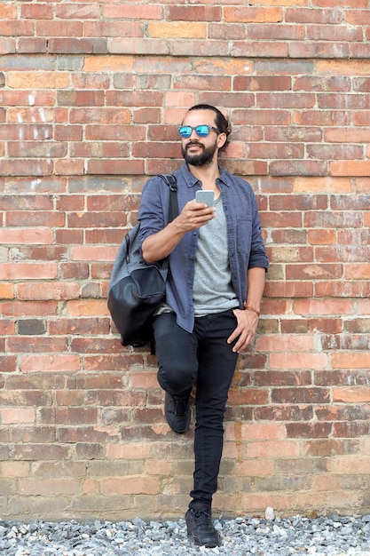 travel, tourism, communication, technology and people concept - happy man with backpack and smartphone over brick wall on street