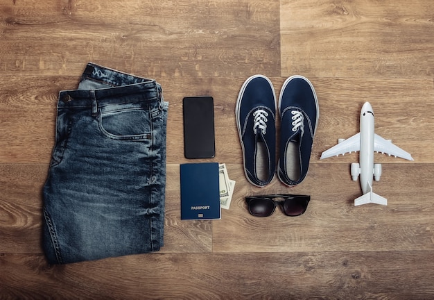 Concetto di viaggio o turismo. jeans, banconote da cento dollari, scarpe da ginnastica, smartphone, passaporto, occhiali da sole, aeroplano su un pavimento di legno. vista dall'alto. lay piatto