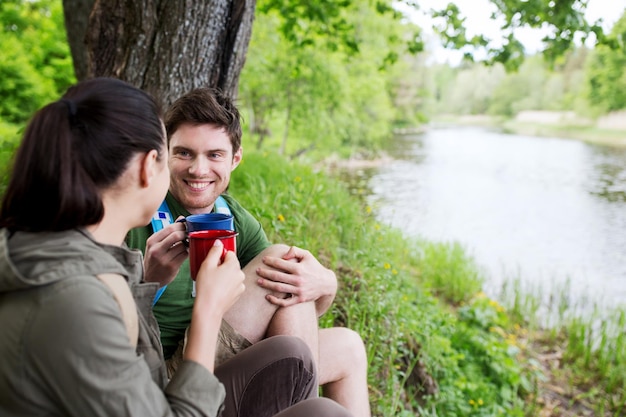 travel, tourism, hike, camping and people concept - happy couple with cups drinking tea in nature on river bank