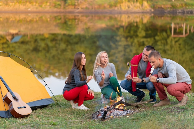 Концепция перемещения, туризма, похода, пикника и людей - группа в составе счастливые друзья жаря сосиски на лагерном костере около озера.