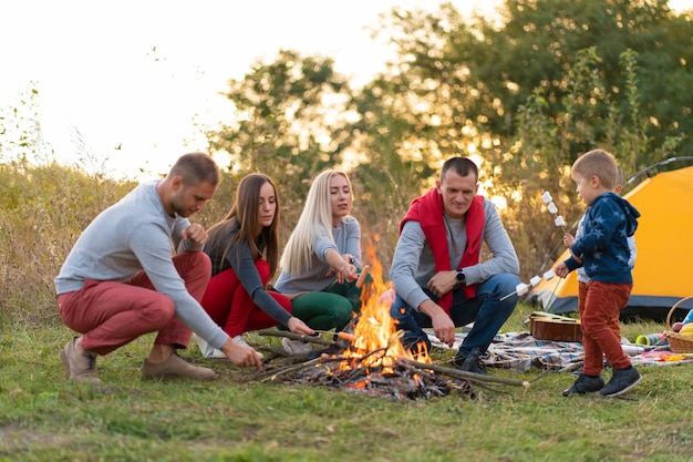 Travel, tourism, hike, picnic and people concept - group of happy friends with kids frying sausages on campfire.