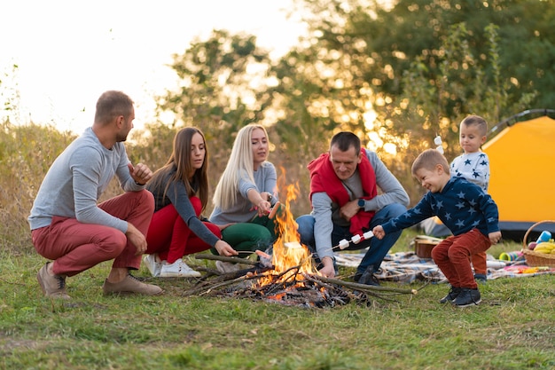 Travel, tourism, hike, picnic and people concept - group of happy friends with kids frying sausages on campfire.