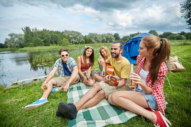 Photo travel, tourism, hike, picnic and people concept - group of happy friends with tent and drinks playing guitar at camping