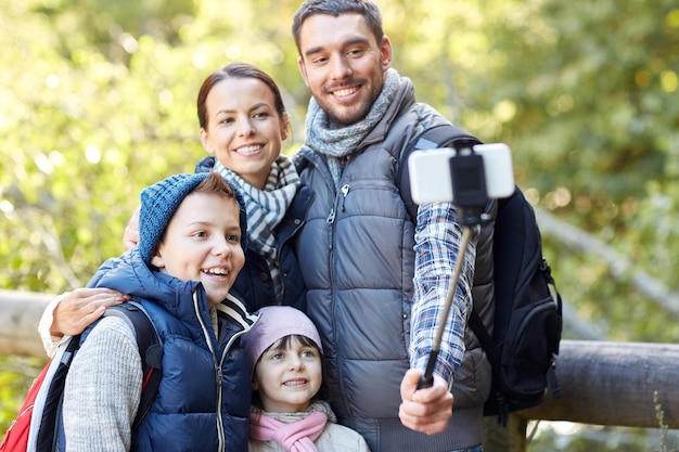 путешествия, туризм, походы, технологии и концепция людей - счастливая семья с рюкзаками, фотографирующая смартфоном и палкой для селфи в лесу