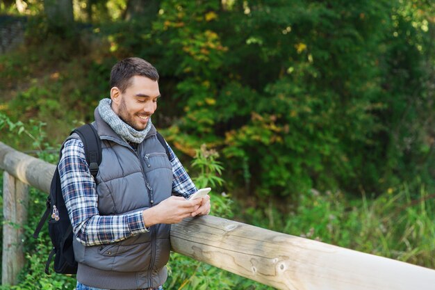 travel, tourism, hike, technology and people concept - happy man with backpack and smartphone texting message outdoors